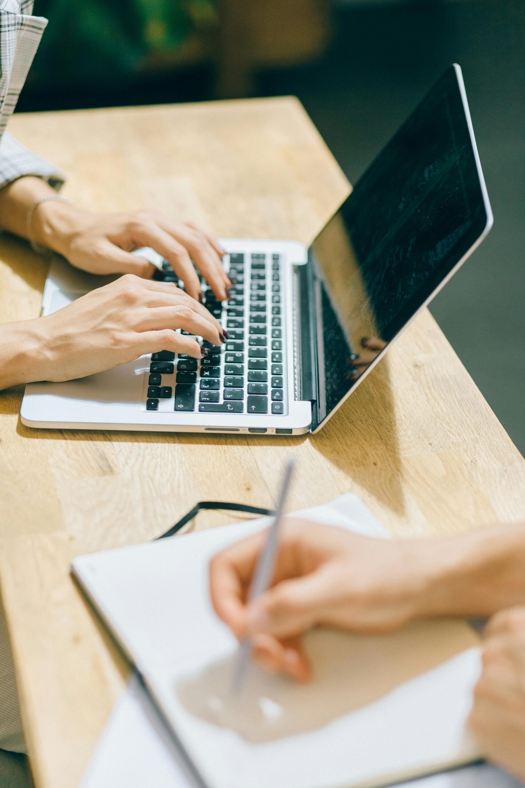 An image of someone typing on a laptop while another person writes in a notebook.